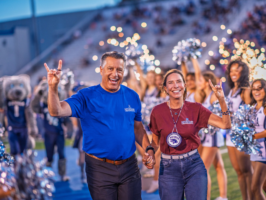 Two individuals walk hand-in-hand across a football field during a sports event, surrounded by cheerleaders with pom-poms. One wears a blue shirt and black pants, the other a maroon shirt and jeans with a large medallion. Both smile and make hand gestures with raised pinky and index fingers. Stadium spectators are visible in the blurred background.