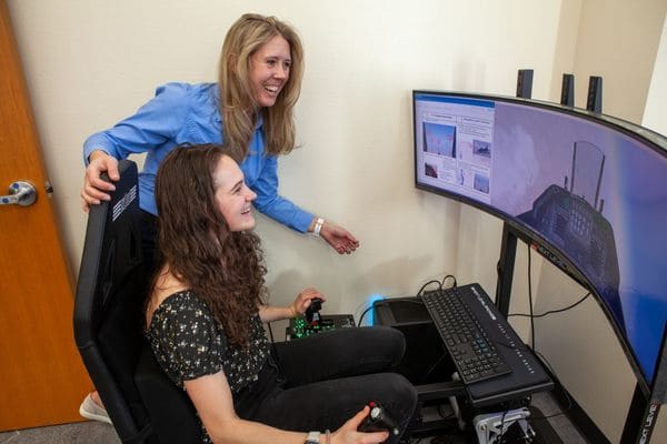 : Student sits at computer with teacher watching over her as they run a flight simulation.