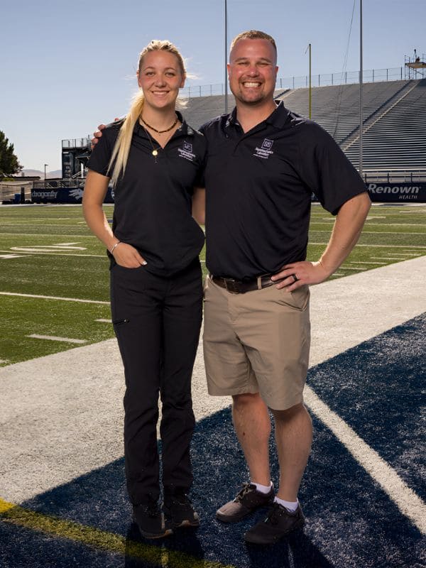 A man and woman stand together on a football field, surrounded by green grass and goalposts in the background.