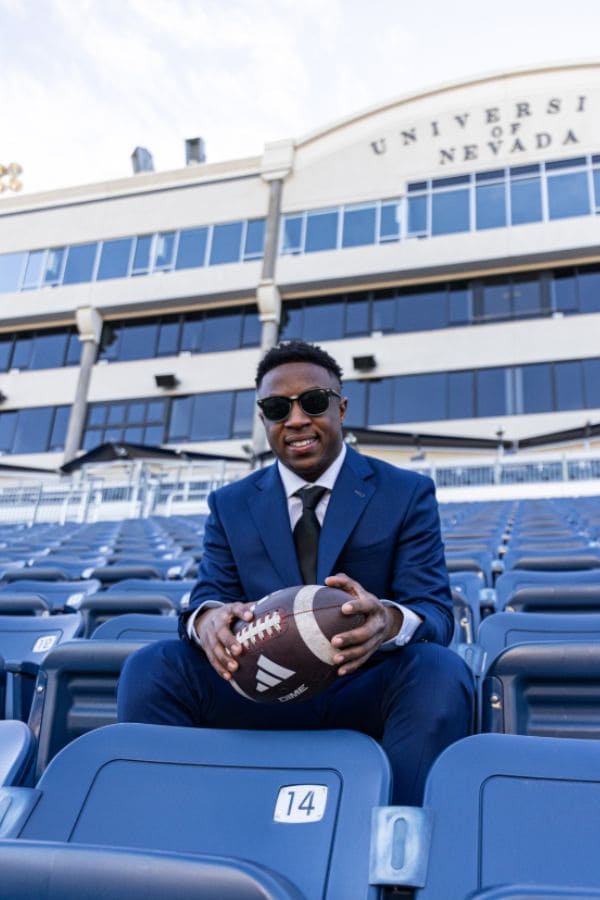 Tori Daffin wears a blue suit and sunglasses as he sits in the stands at Mackay Stadium while holding a football. A sign above reads "University of Nevada."