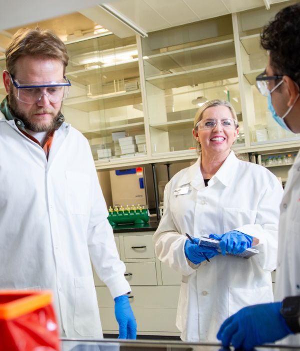 Three scientists in a lab wearing white coats, safety goggles, and gloves. They are engaged in discussion 