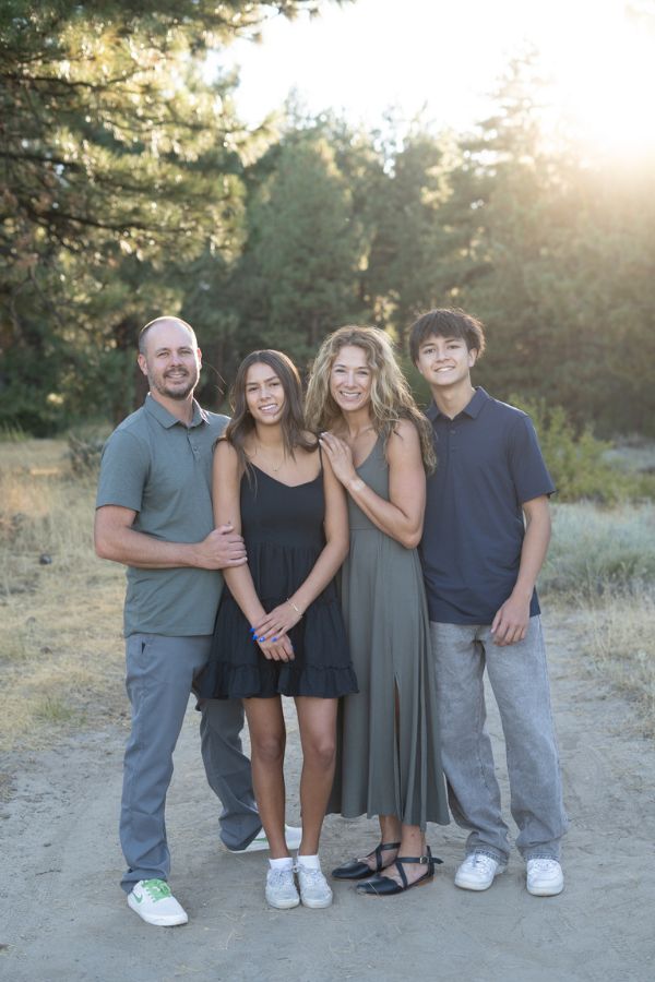 A family of four stands closely in an outdoor setting with tall trees and sunlight streaming through. They are smiling, creating a warm atmosphere.