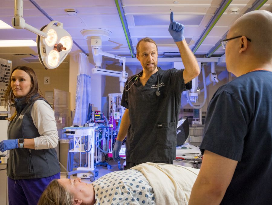Medical team in scrubs discussing a patient's treatment in a hospital room. One doctor gestures upwards, conveying a sense of urgency.