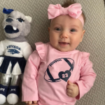 A smiling baby in a pink outfit with a football design lies next to a wolf mascot toy wearing a cheerleader uniform. The baby wears a pink bow.