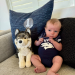 A baby wearing a navy onesie sits on a couch beside a plush husky toy. Behind them, a blue pillow adds a cozy background. The scene is cute and calm.