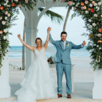 A joyful bride in a white gown and groom in a blue suit lift their arms under a floral arch, celebrating on a beach with palm trees and ocean in the background.