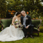 A bride and groom sit closely on a vintage green couch in a garden, gazing at each other lovingly. The bride holds a bouquet, and both are smiling.