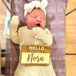 A newborn baby lies peacefully on a patterned blanket labeled "Nora Jean." The baby wears a light onesie and a white bow headband, exuding warmth and joy.