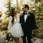 A joyful couple, dressed in wedding attire, stands outdoors. The bride holds a bouquet and wears sunglasses, while both share a big smile, surrounded by greenery.