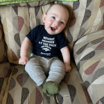 A smiling baby in a black t-shirt sits on a patterned, beige and brown sofa. The baby looks happy and relaxed with an engaging demeanor.