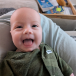 A smiling baby in a green onesie lies on a pillow, with books and a toy in the background, conveying a joyful and playful mood.