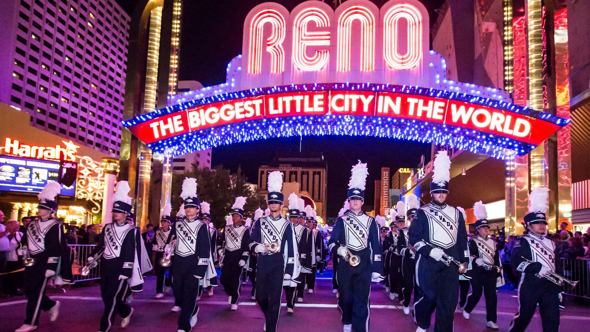 The Wolf Pack Marching Band performs at night under the illuminated Reno Arch, with vibrant outfits and instruments.