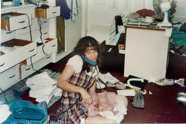 Christ Jerz has a smile and sits on a cluttered floor, holding a phone. An iron and scattered clothes surround her in a messy room. 