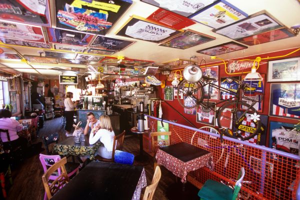 A vibrant, eclectic caf&eacute; with colorful posters on the ceiling and walls. Patrons sit at mismatched tables. A bicycle hangs on a red wall, creating a cozy, artistic vibe. 