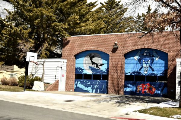 Brick building with two blue garage doors featuring murals of a silhouette and a roaring animal. A basketball hoop stands on the left of the building. 