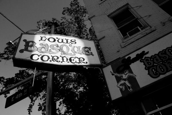 Black-and-white photo of the &ldquo;Louis' Basque Corner&rdquo; sign on a street corner. The vintage sign contrasts with the tree shadows and a brick building. 