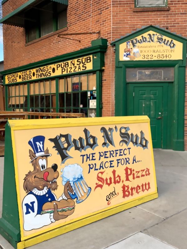 Pub exterior with a rustic brick fa&ccedil;ade and green door. A colorful sign featuring a cartoon wolf holds a beer and a sub, advertising &ldquo;Pub N&rsquo; Sub&rdquo; for subs, pizza, and brews