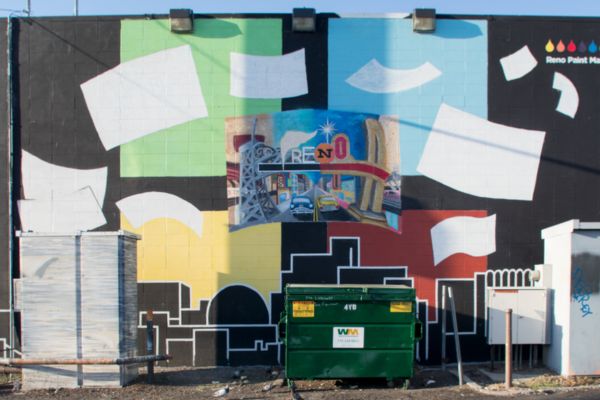 Colorful mural on a wall features abstract shapes with a central image of Reno, including iconic landmarks. A green dumpster sits in front of the mural. 