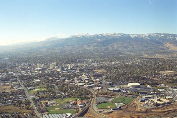 Aerial view of a cityscape with a cluster of buildings in the center, surrounded by greenery. Snow-capped mountains and a blue sky provide a scenic backdrop.