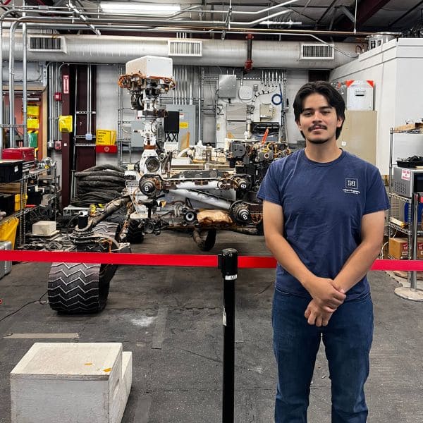 A man in a blue t-shirt stands in front of a Mars rover model inside a workshop filled with machinery and equipment, conveying a sense of innovation and exploration.
