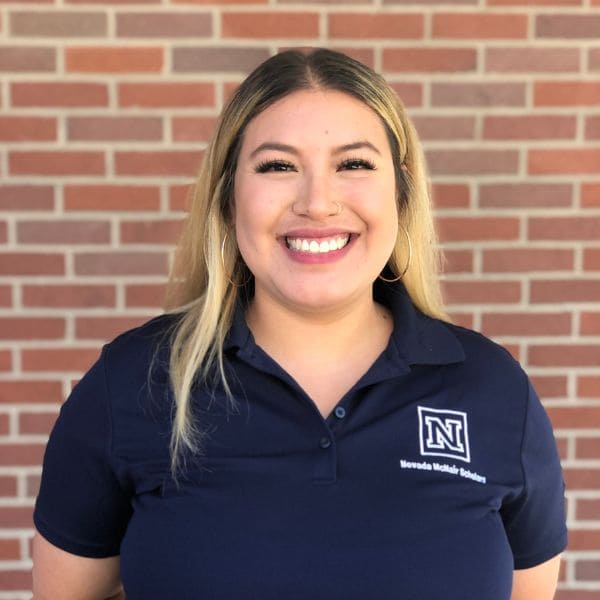 Smiling woman with long hair and hoop earrings stands against a brick wall. She wears a navy shirt with an emblem and appears confident and approachable.