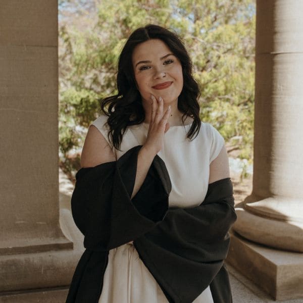 Smiling woman with dark hair and white dress, draped in a black graduation gown. She’s standing in a columned outdoor setting.