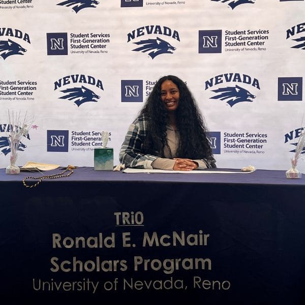 A woman with long hair smiles while seated at a table with a "TRiO Ronald E. McNair" banner. Behind her is a University of Nevada, Reno backdrop.