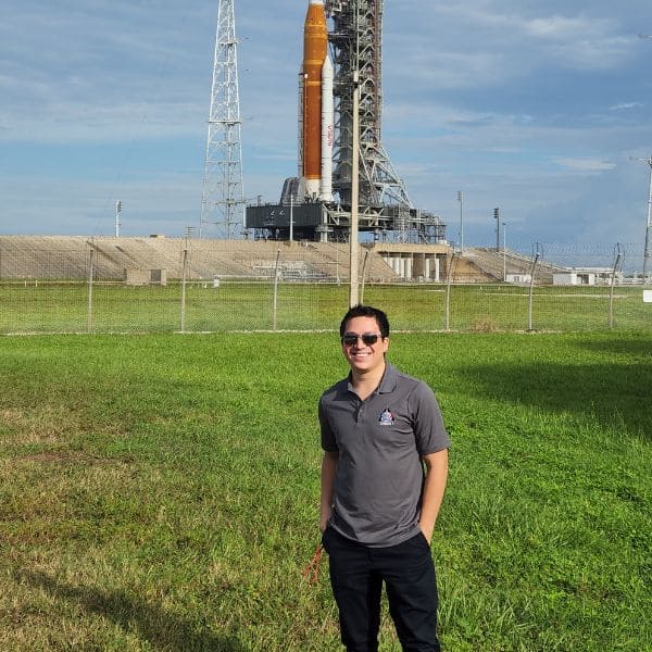 Manuel smiles in front of a space launch pad under a clear sky. 