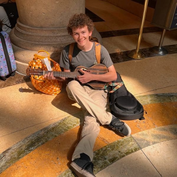 A young person with curly hair smiles while sitting on a polished floor, playing a ukelele. They are by a large column, next to a colorful bag. 