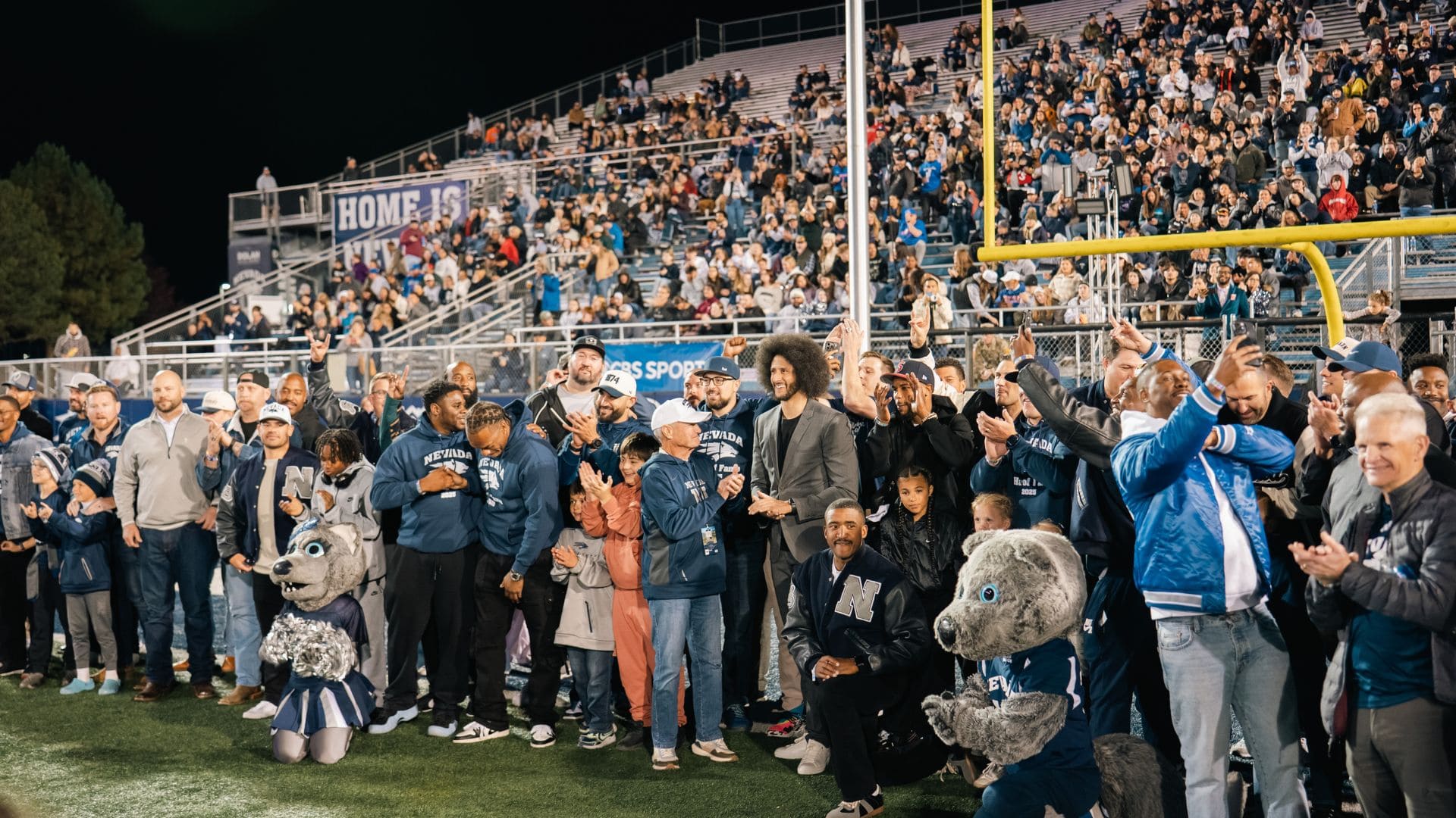 Members of the 2010 Nevada Football team, including mascots and fans in sports attire, stands on the Mackay football field at night. The crowd in the background applauds enthusiastically. 