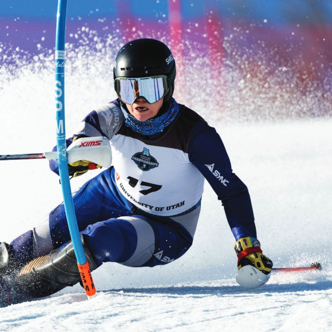 Skier in mid-action, leaning sharply as they navigate a slope, snow spraying around. The skier wears a black helmet, mirrored goggles, and a numbered bib. 