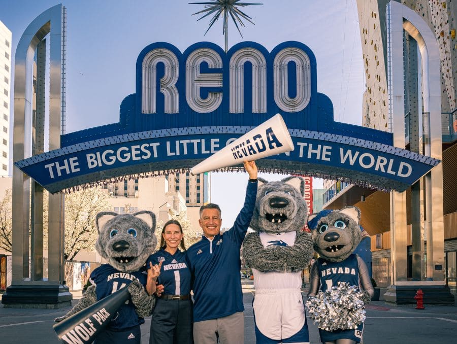 A group poses under the iconic Reno arch. President Sandoval (right) and First Lady Lauralyn Sandoval stand while holding a megaphone with the word Nevada on it, flanked by with the Nevada Wolf Pack mascots holding Nevada and Wolf Pack signs. 