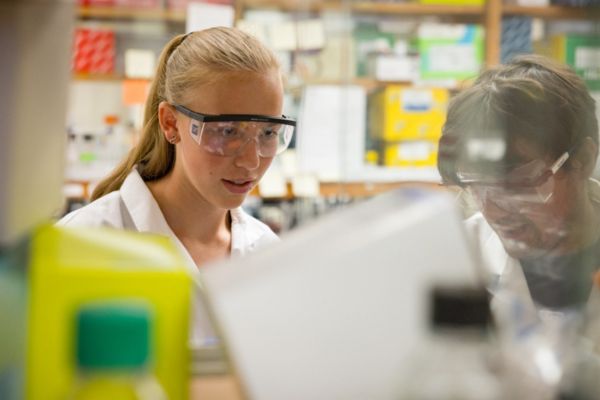 Two people wearing lab coats and safety goggles work intently in a busy laboratory, surrounded by colorful equipment and materials.