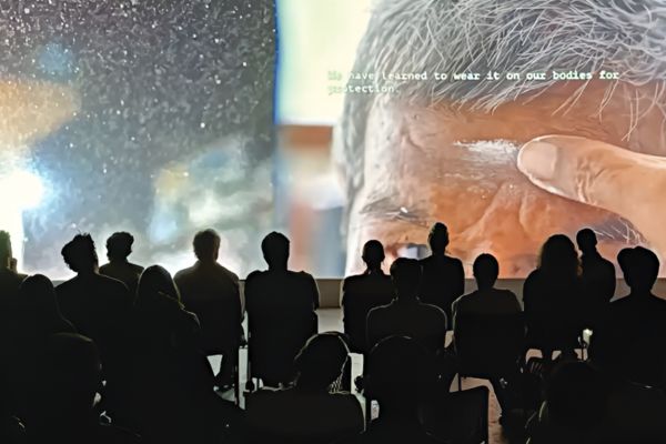 A group of people sits in silhouette watching a large screen. The screen shows a close-up of a person&rsquo;s face with text overlayed. 