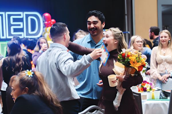 A joyful group at a celebration; a woman holding a bouquet hugs a man with a bright smile. Balloons and neon sign in the background add festivity.