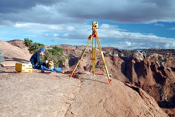 A person sits on a rocky desert ledge beside surveying equipment mounted on a tripod, overlooking a rugged canyon landscape under a partly cloudy sky. Nearby are a yellow equipment case and scattered tools, with layered red and brown rock formations stretching into the distance.