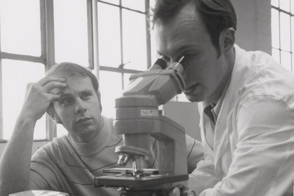 Two men in a lab, one in a sweater looking thoughtful, the other in a lab coat examining a microscope.