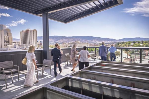 A group of people enjoy a cityscape view from a modern rooftop, under a pergola, with mountains in the distance. The scene is bright and tranquil.