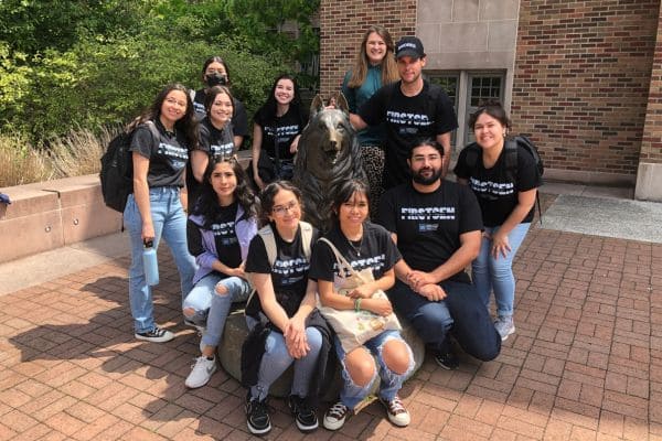 A group of smiling people wearing matching black T-shirts poses around a bronze wolf statue on a brick path. 