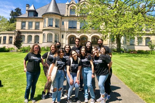 A group of twelve smiling people wearing matching black shirts stands outdoors on a sunny day, in front of a historic stone building with arched windows, surrounded by green grass and trees. 