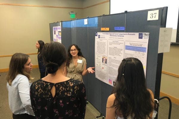 A woman presents research on a poster board to three attentive attendees. They stand in a room with similar poster displays in the background.