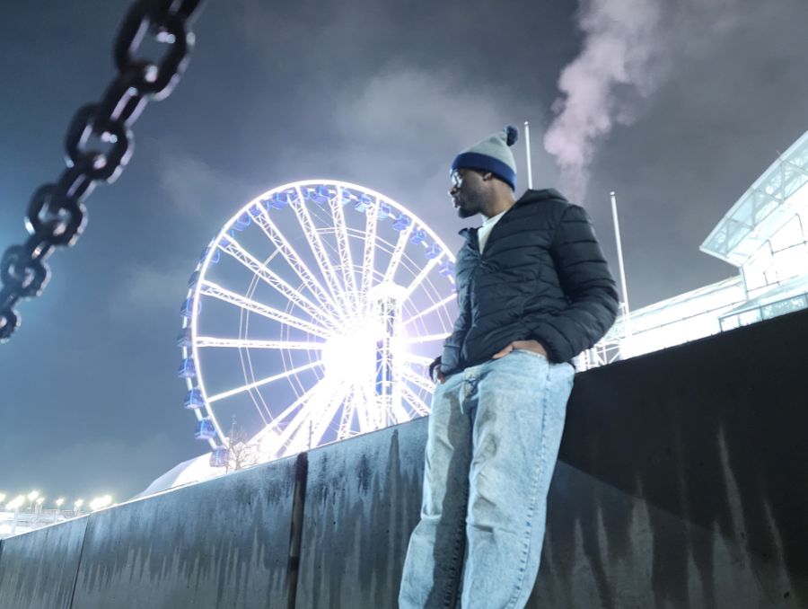 Man in winter attire leans against a railing at night, gazing at an illuminated Ferris wheel in the background. The scene is calm and atmospheric.