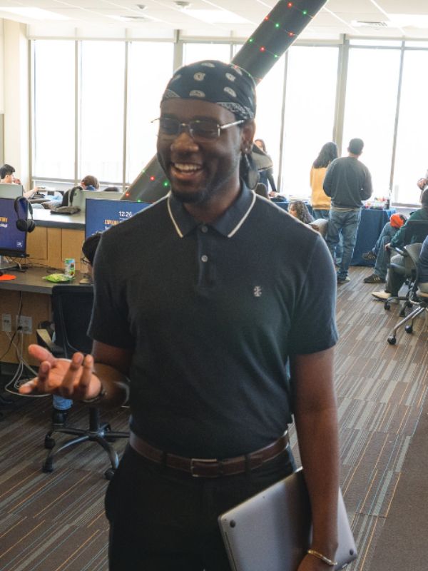A person in a black polo and bandana smiles while holding a laptop in an office. Others work at computers, and sunlight streams through large windows.