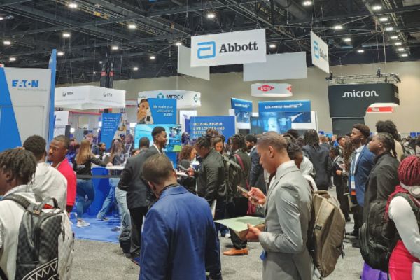: A busy career fair scene with diverse attendees engaging at booths. Prominent signage includes Abbott and Micron, indicating a corporate environment