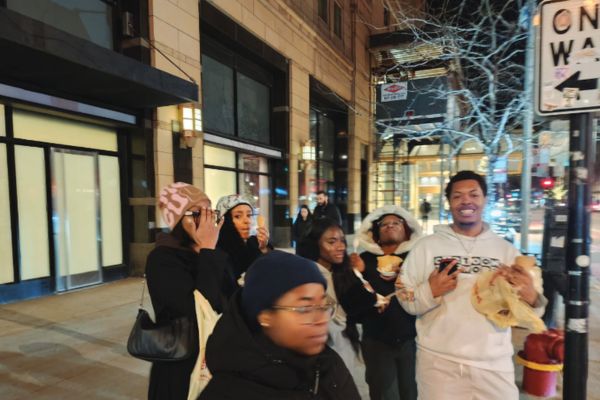 A group of six people walk along a city street at night. They appear cheerful, wearing winter clothing and holding food. The street is lined with lit shops.