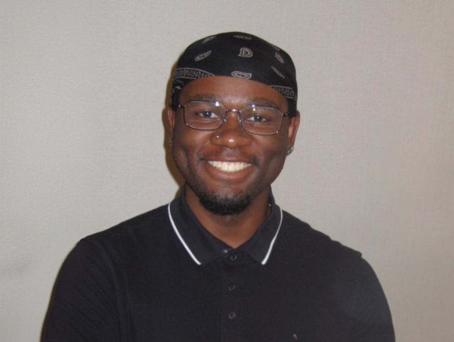 Smiling person wearing glasses, a black bandana, and a dark collared shirt against a neutral background, conveying a friendly and cheerful mood.