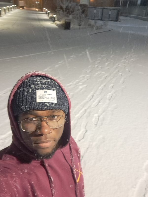 A person in a maroon hoodie and knit hat stands on a snow-covered path at night. Snowflakes fall gently, with footprints trailing behind them.