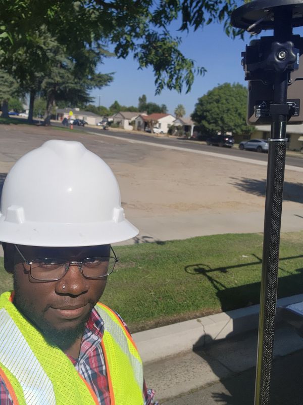 A person wearing a white hard hat and safety vest stands outdoors. A sunny, residential street and trees are in the background.