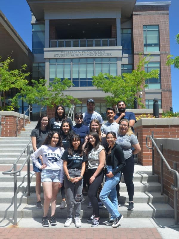 A diverse group of smiling students stands on steps outside the “William N. Pennington Student Achievement Center,” with green trees in the background.