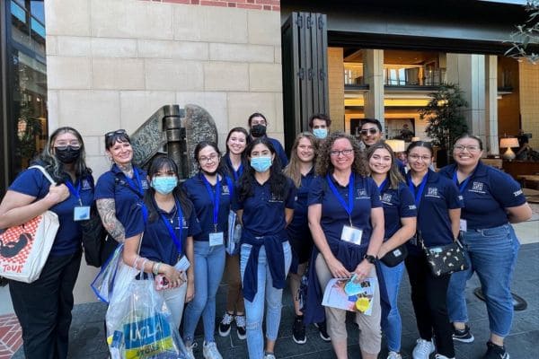 A group of fourteen people, mostly women, in matching navy shirts and lanyards, gathered outside a modern building. They are smiling and appear cheerful.